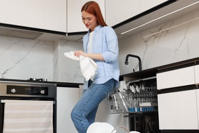 Photo of Beautiful woman wiping plate near open dishwasher in kitchen, low angle view