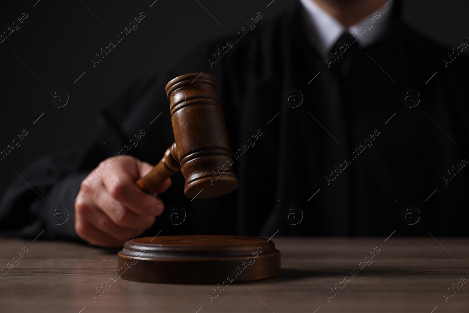 Photo of Judge with gavel sitting at wooden table against black background, closeup
