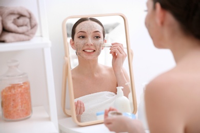 Photo of Woman applying scrub onto face in front of mirror at home