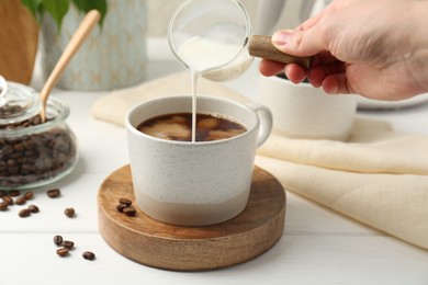 Photo of Woman pouring milk into cup with coffee at white wooden table, closeup
