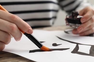Photo of Calligraphy. Woman with brush writing hieroglyphs on paper at table, closeup