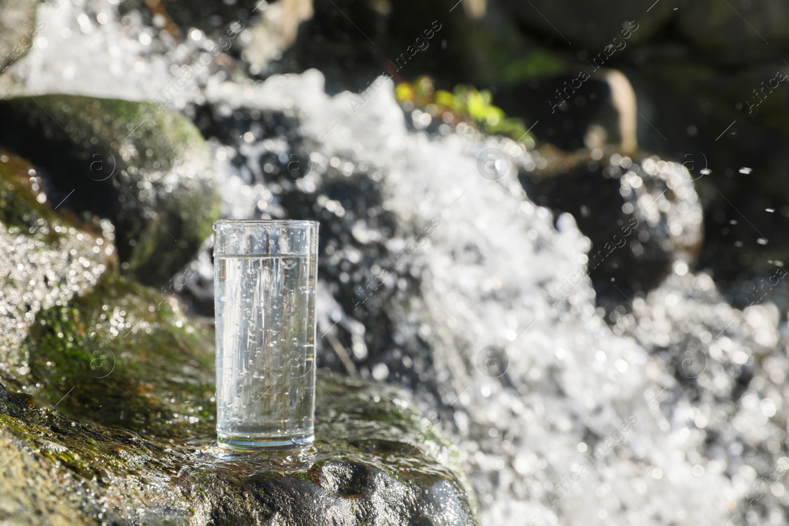 Photo of Wet glass of water on rocks near flowing stream outdoors, space for text