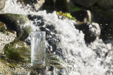 Photo of Wet glass of water on rocks near flowing stream outdoors, space for text