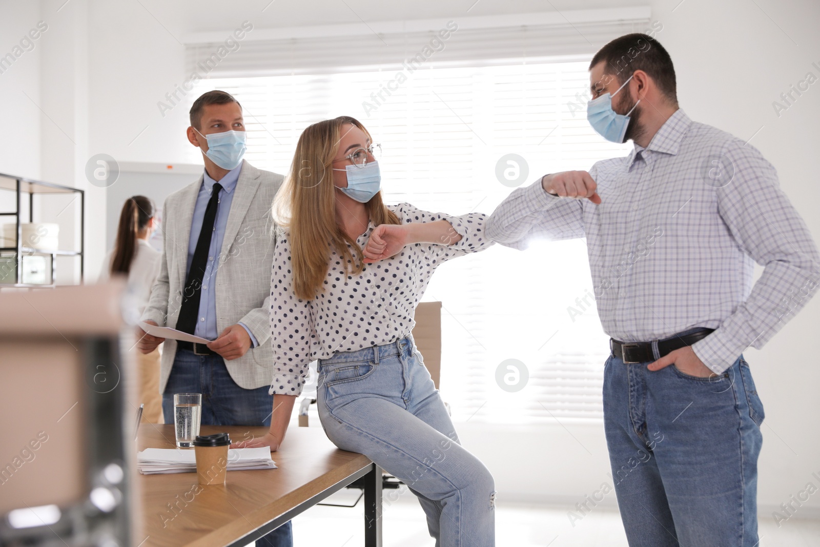 Photo of Coworkers with protective masks making elbow bump in office. Informal greeting during COVID-19 pandemic