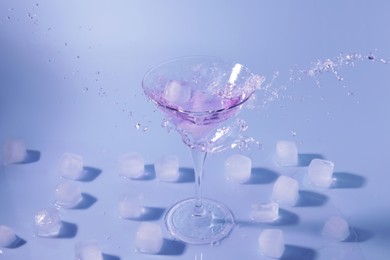 Photo of Fresh cocktail splashing out of martini glass near ice cubes on light blue background