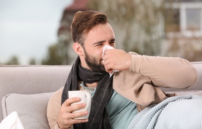 Photo of Ill man with cup of hot tea for cough on sofa at home