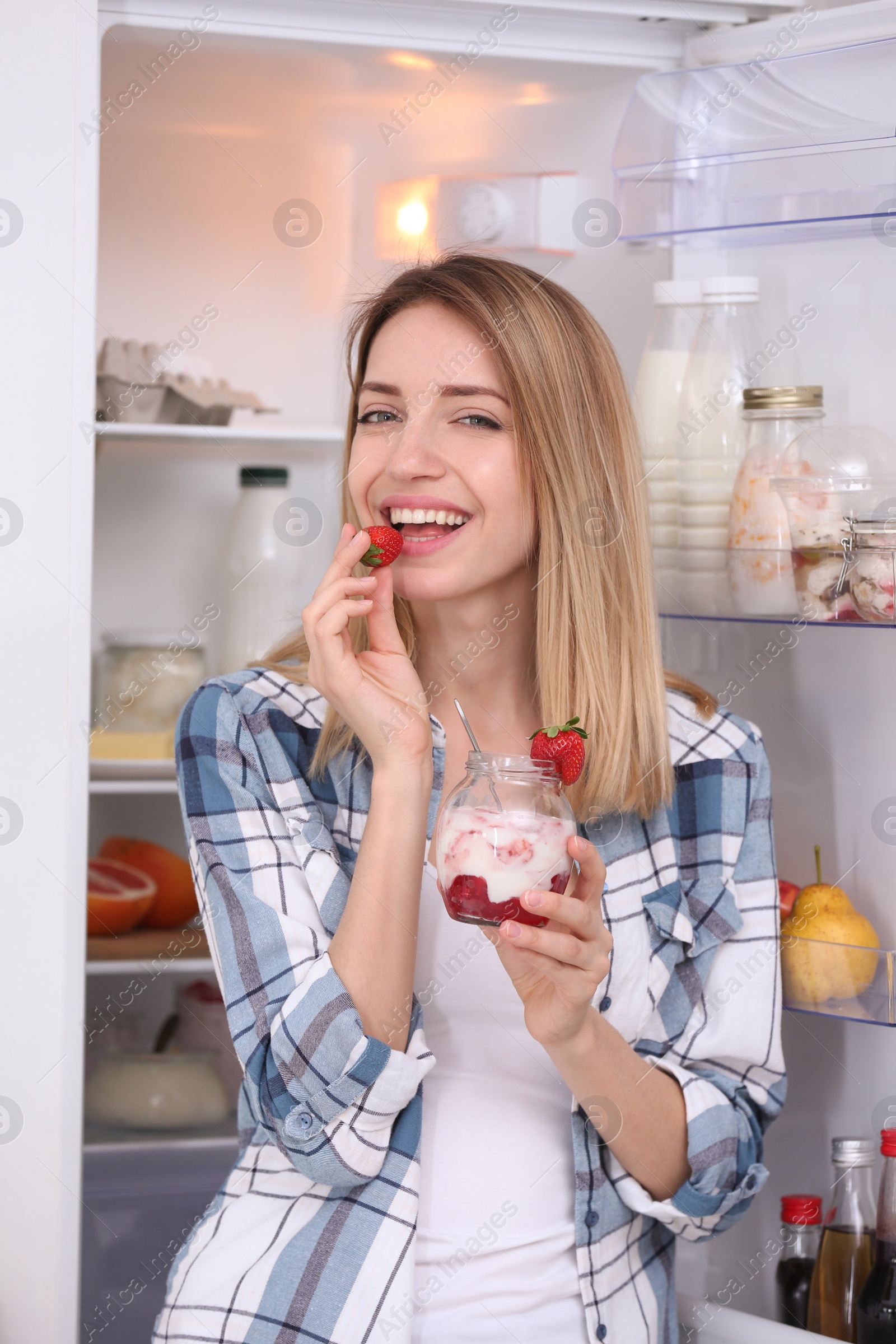 Photo of Young attractive woman eating tasty yogurt near fridge