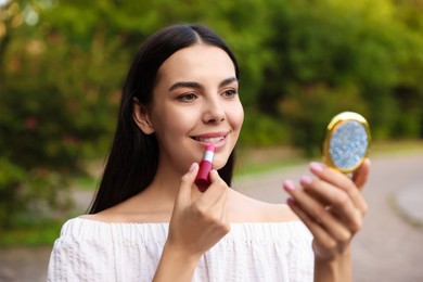 Photo of Beautiful young woman with cosmetic pocket mirror applying lipstick outdoors