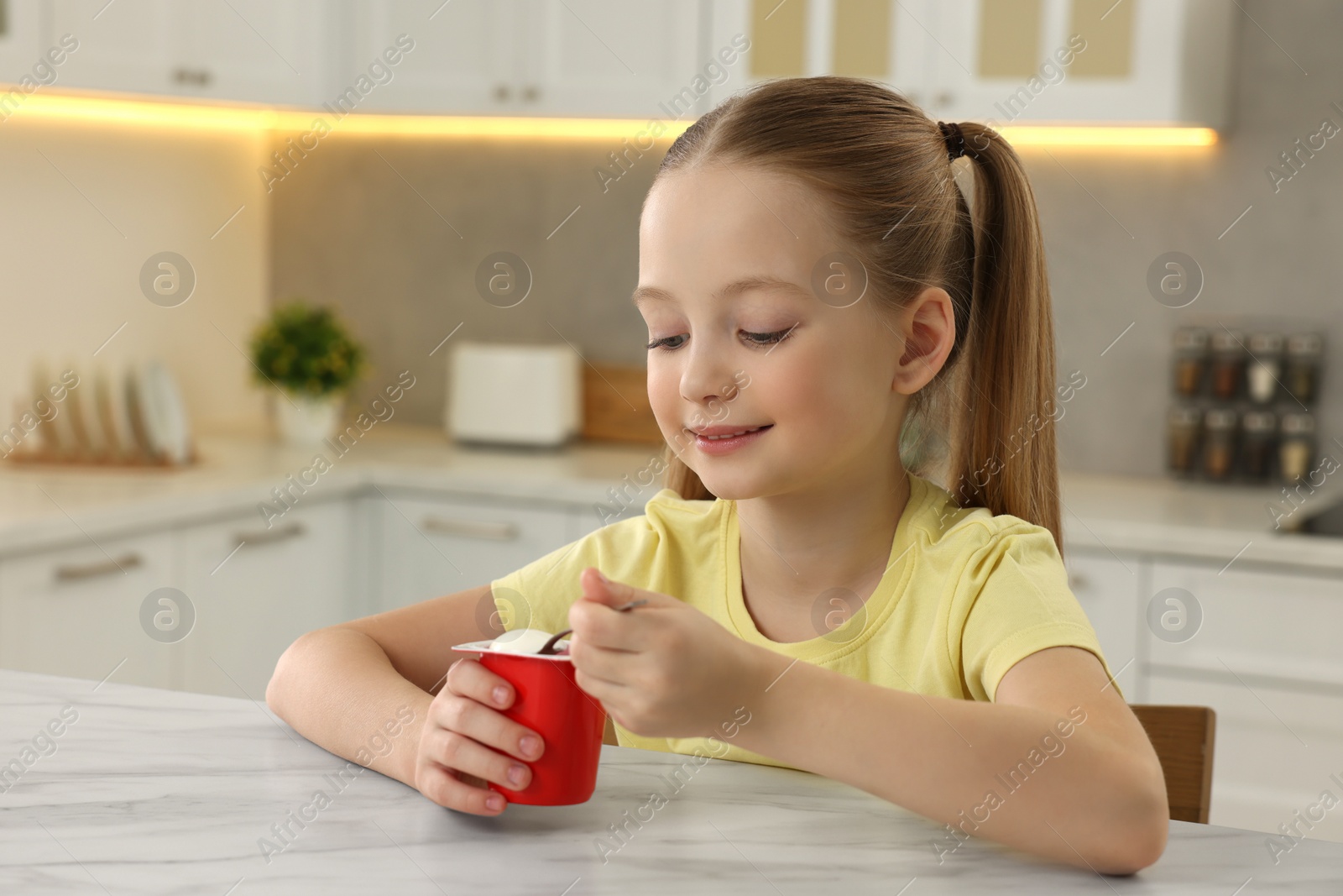 Photo of Cute little girl with tasty yogurt at white marble table in kitchen