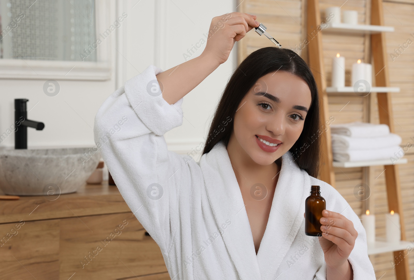 Photo of Happy young woman with bottle applying essential oil onto hair roots in bathroom