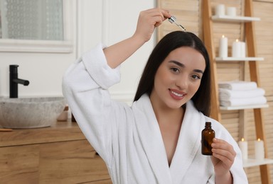 Photo of Happy young woman with bottle applying essential oil onto hair roots in bathroom