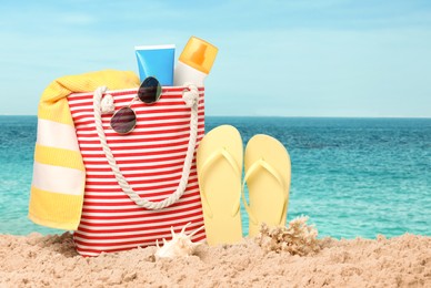 Stylish bag with different accessories on sandy beach near ocean