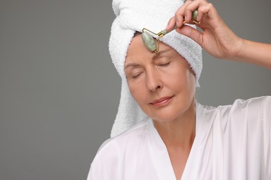 Woman massaging her face with jade roller on grey background, space for text