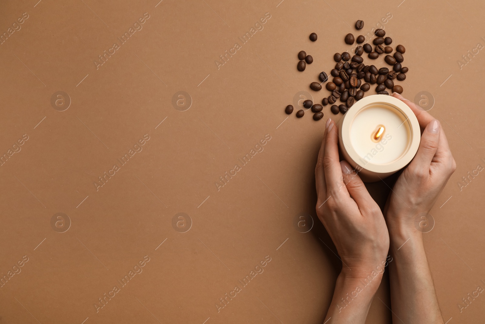 Photo of Woman with burning soy candle and coffee beans on brown background, top view. Space for text