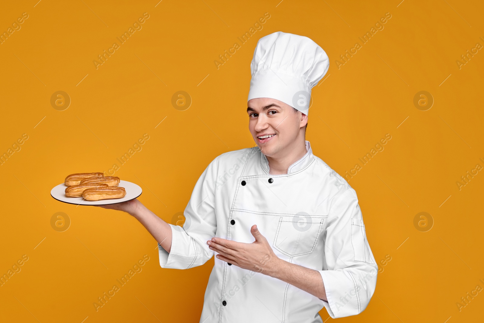 Photo of Portrait of happy confectioner in uniform holding plate with eclairs on orange background