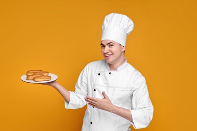 Photo of Portrait of happy confectioner in uniform holding plate with eclairs on orange background