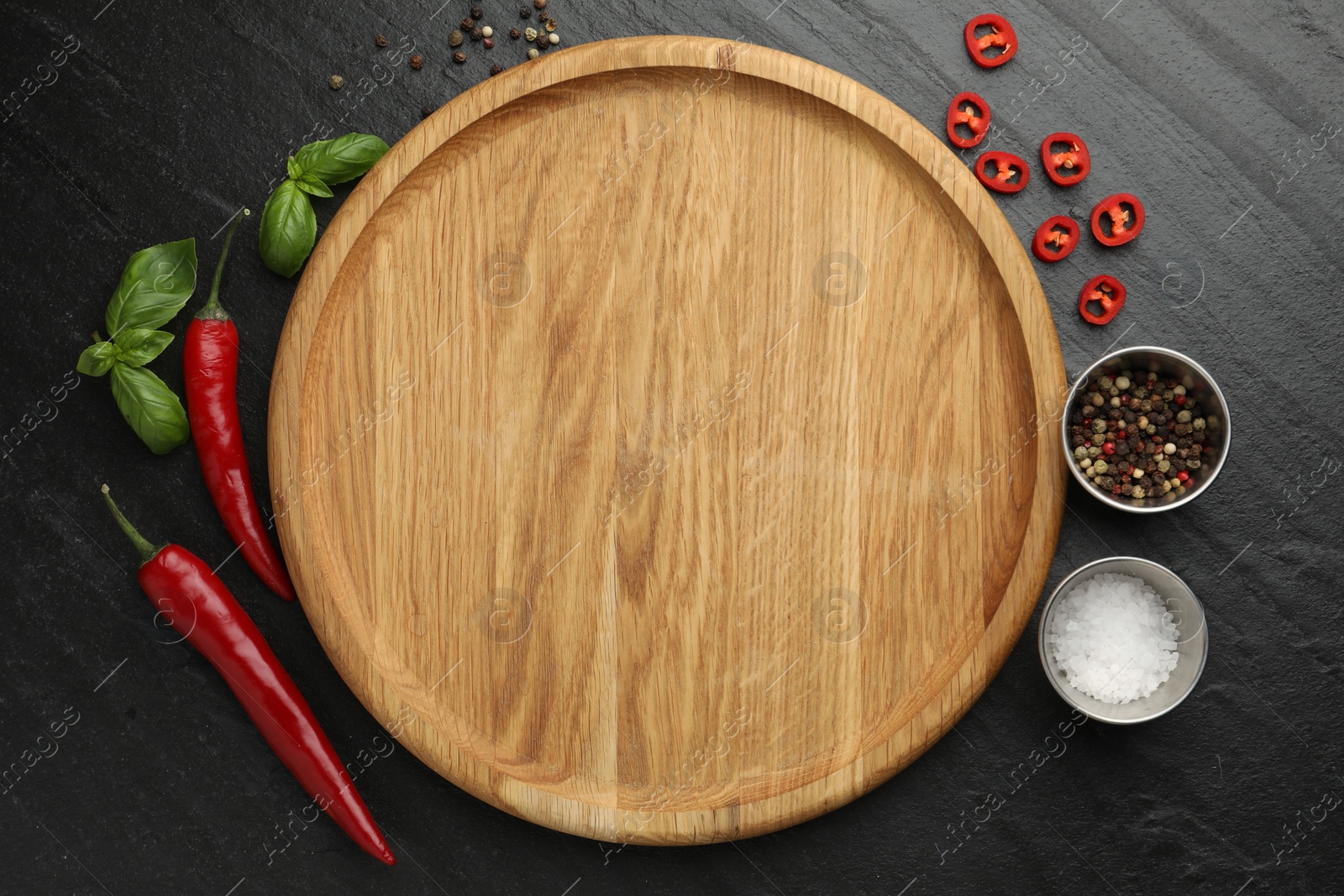 Photo of Cutting board, salt, spices, basil and chili peppers on black textured table, flat lay. Space for text