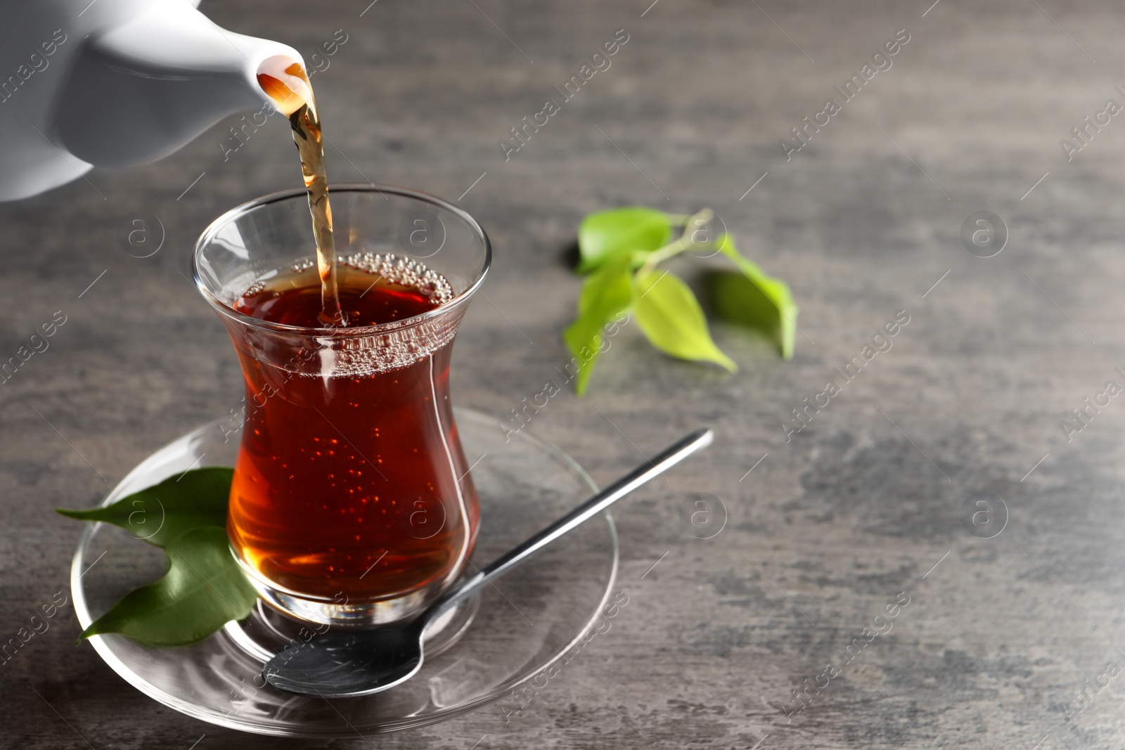 Photo of Pouring traditional Turkish tea from pot into glass on grey table, space for text