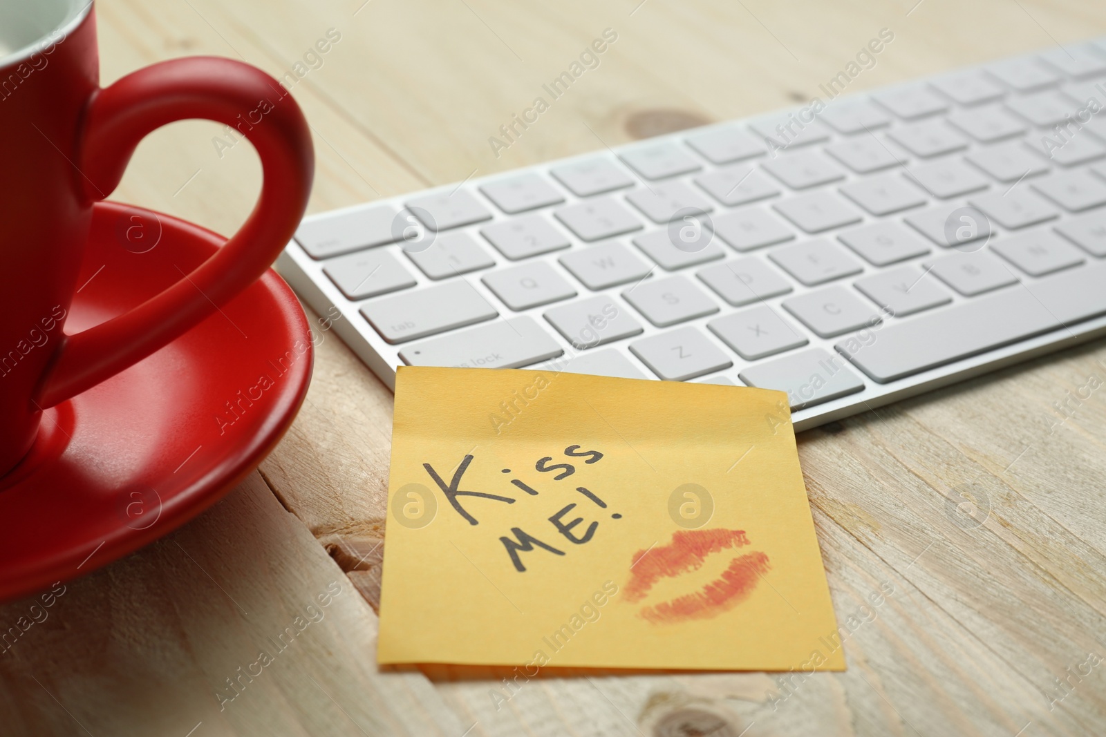 Photo of Sticky note with phrase Kiss Me, lipstick mark, cup of coffee and keyboard on wooden table, closeup