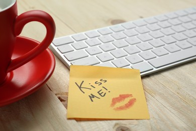 Photo of Sticky note with phrase Kiss Me, lipstick mark, cup of coffee and keyboard on wooden table, closeup