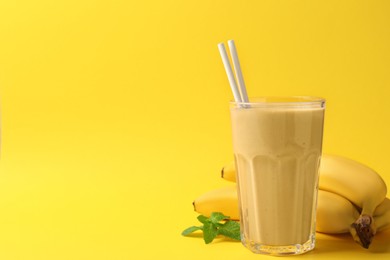 Glass of tasty banana smoothie with straws, fresh fruits and mint on yellow background. Space for text