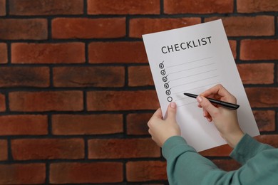 Woman filling Checklist against brick wall, closeup. Space for text