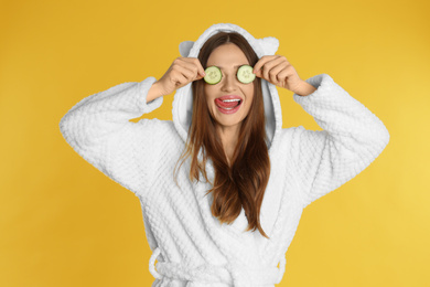 Photo of Young woman in bathrobe with cucumber slices on yellow background