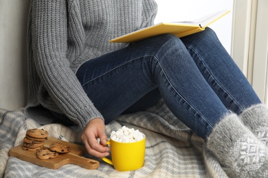 Photo of Woman with cup of hot cocoa reading book on window sill, closeup. Winter drink