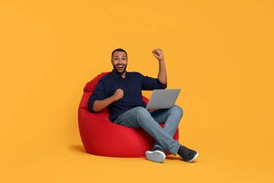 Happy young man with laptop sitting on beanbag chair against yellow background