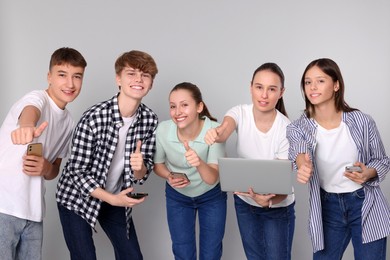 Photo of Group of happy teenagers with smartphones and laptop showing thumbs up on light grey background