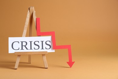 Photo of Wooden easel with word Crisis and descending red arrow on brown background, space for text