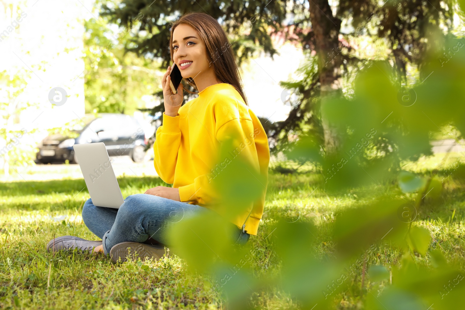 Image of Happy young woman with laptop talking on phone in park