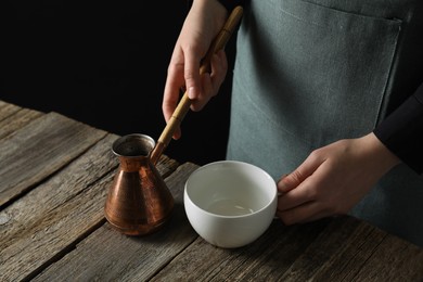 Photo of Turkish coffee. Woman with cezve and cup at wooden table against black background, closeup