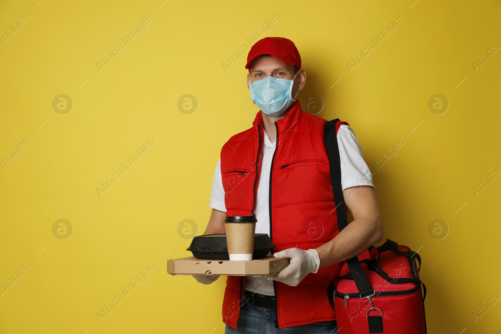 Photo of Courier in protective gloves and mask holding order on yellow background. Food delivery service during coronavirus quarantine