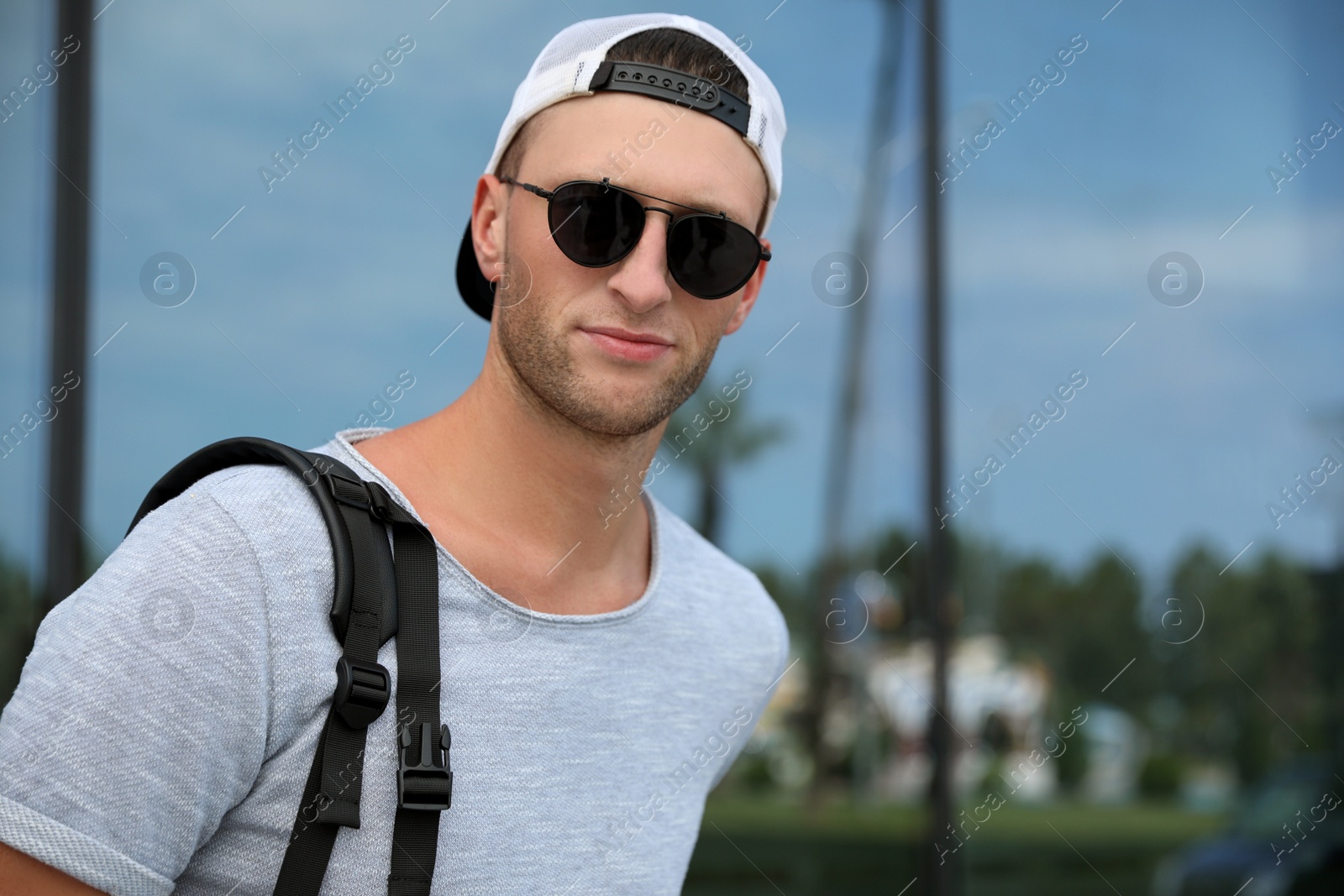 Photo of Handsome young man with stylish sunglasses and backpack near reflection surface outdoors, space for text