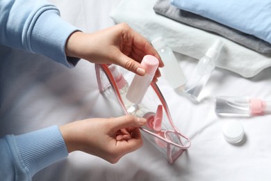 Photo of Cosmetic travel kit. Woman putting small bottle with personal care product into plastic bag on bed, closeup