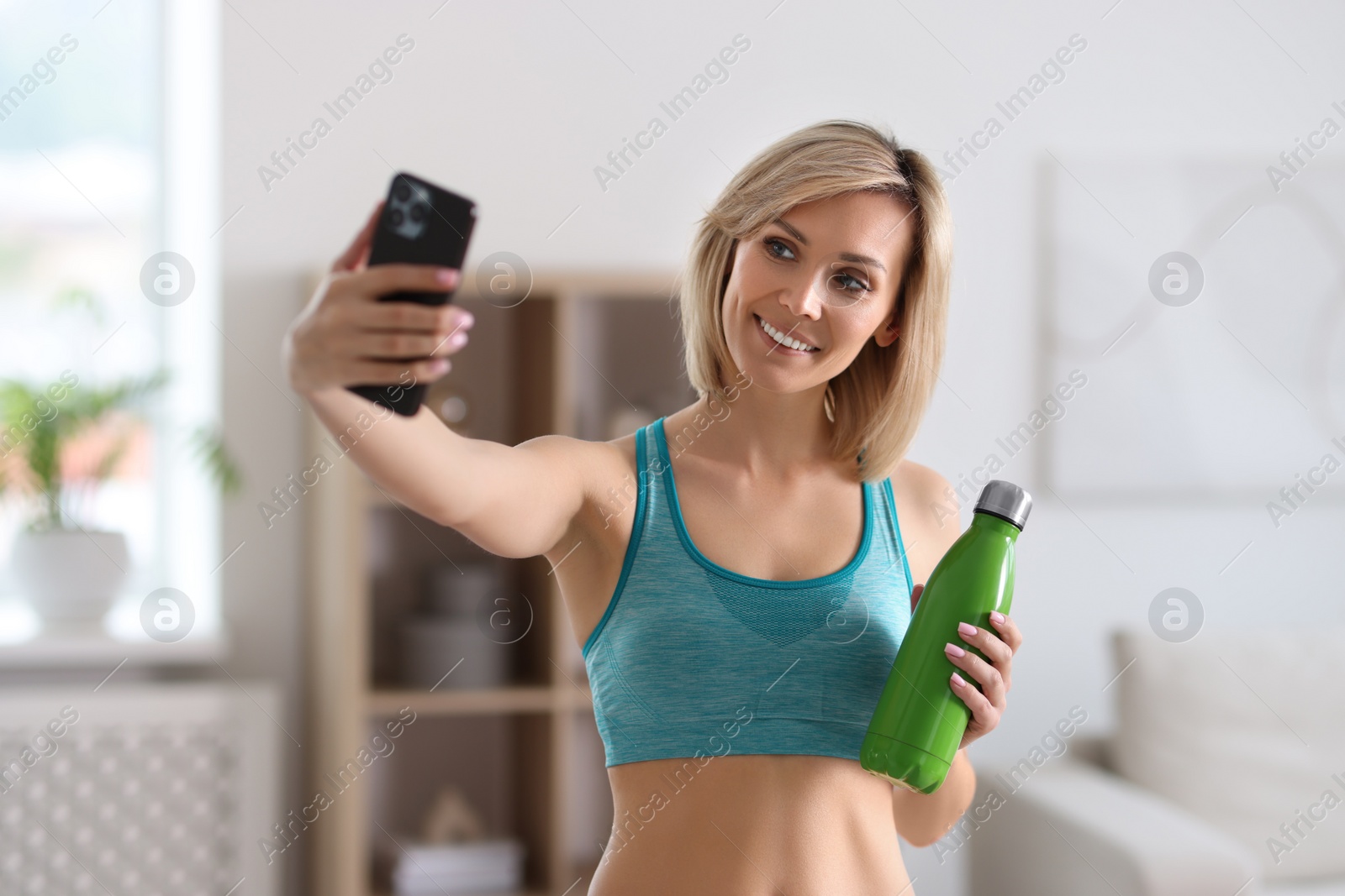 Photo of Smiling sports blogger with bottle streaming online fitness lesson with smartphone at home