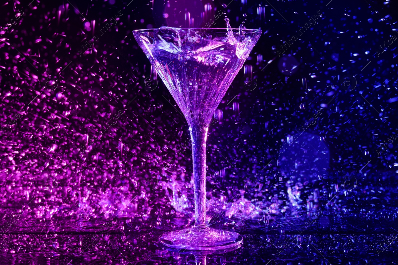 Photo of Glass of martini and splashes in neon lights on dark blue background, bokeh effect