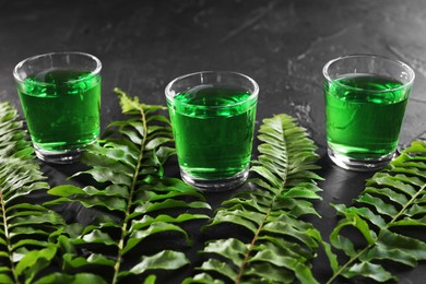 Photo of Absinthe in shot glasses and green leaves on black table, closeup. Alcoholic drink