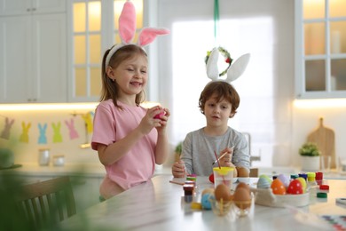 Photo of Easter celebration. Cute children with bunny ears painting eggs at white table in kitchen