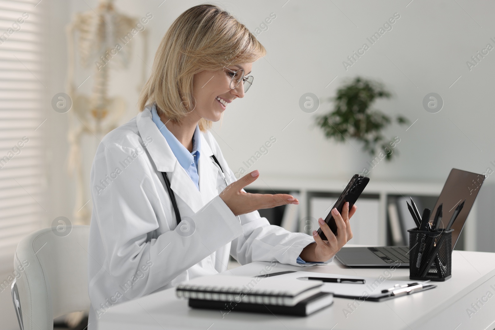 Photo of Smiling doctor with smartphone having online consultation at table in office