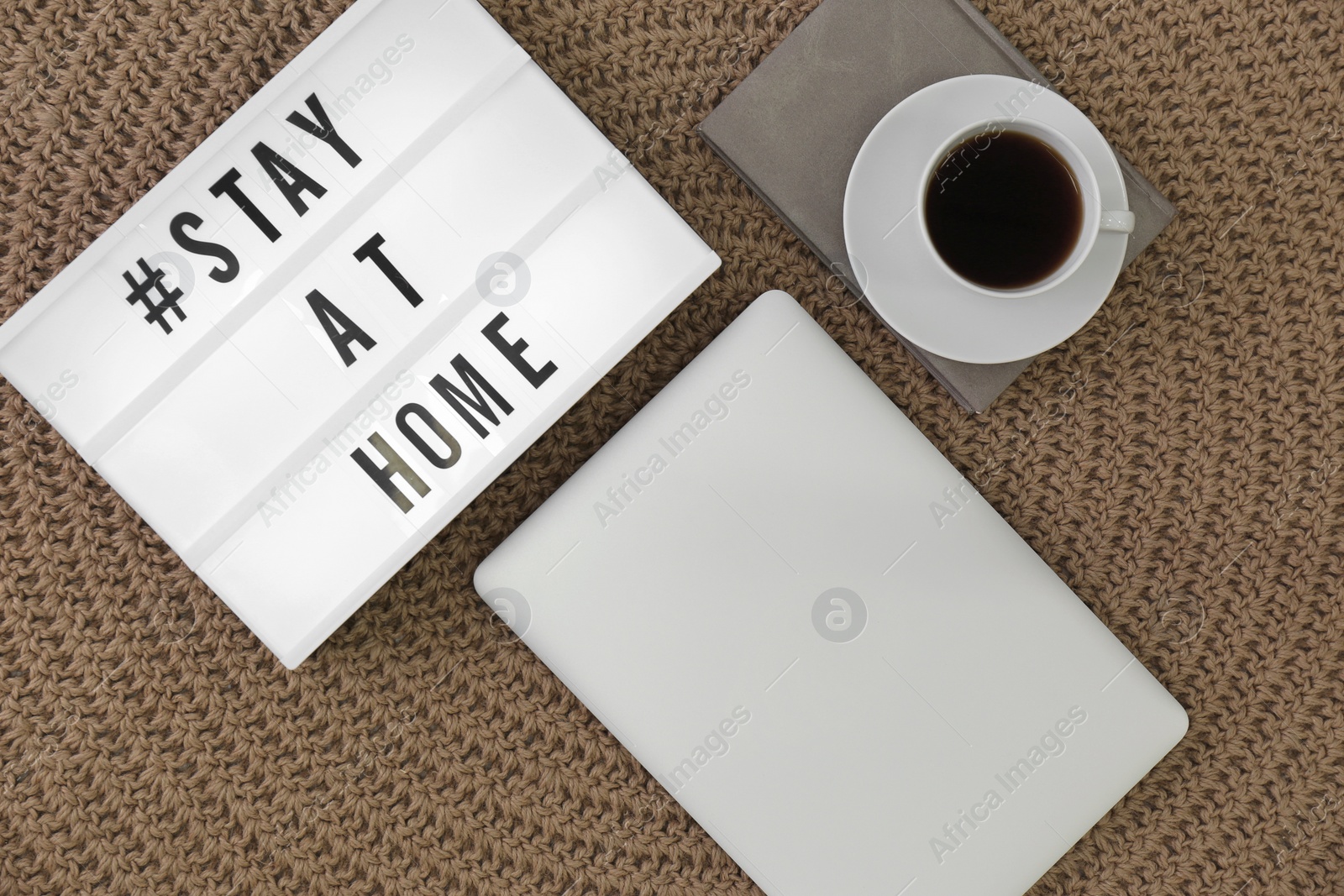 Photo of Laptop, cup of coffee and lightbox with hashtag STAY AT HOME on knitted rug, flat lay. Message to promote self-isolation during COVID‑19 pandemic