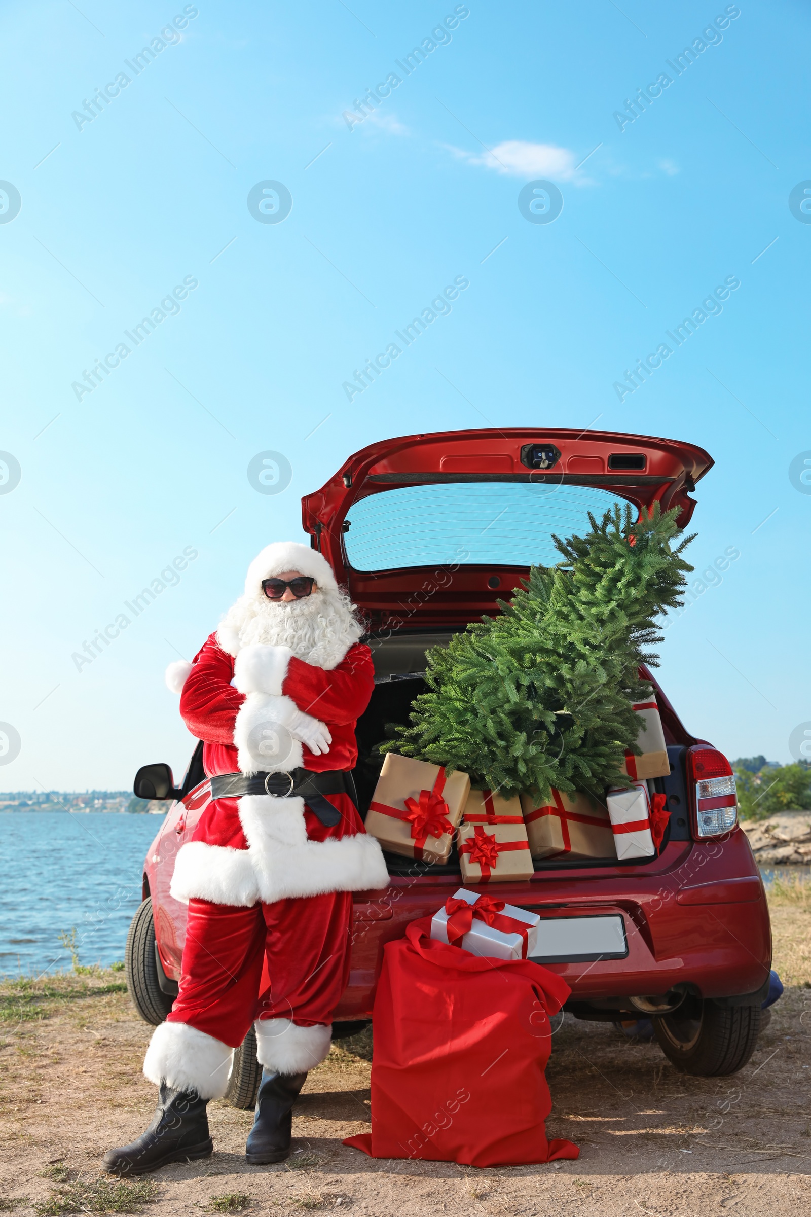 Photo of Authentic Santa Claus near red car with gift boxes and Christmas tree on beach