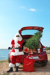 Authentic Santa Claus near red car with gift boxes and Christmas tree on beach