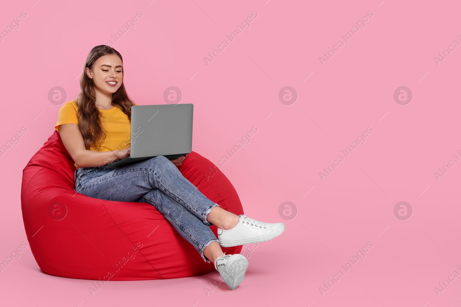 Photo of Happy woman with laptop sitting on beanbag chair against pink background. Space for text