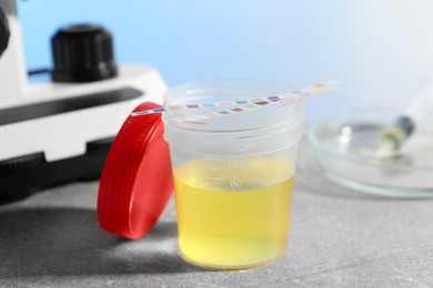 Photo of Container with urine sample for analysis and test strips on grey table in laboratory, closeup