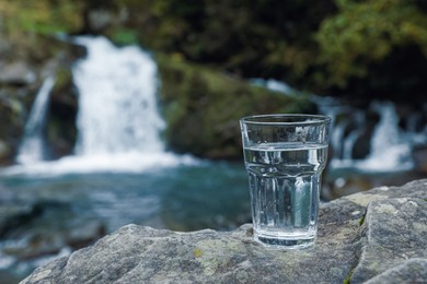 Glass of fresh water on stone near waterfall outdoors. Space for text