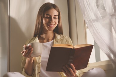 Image of Beautiful young woman reading book near window at home