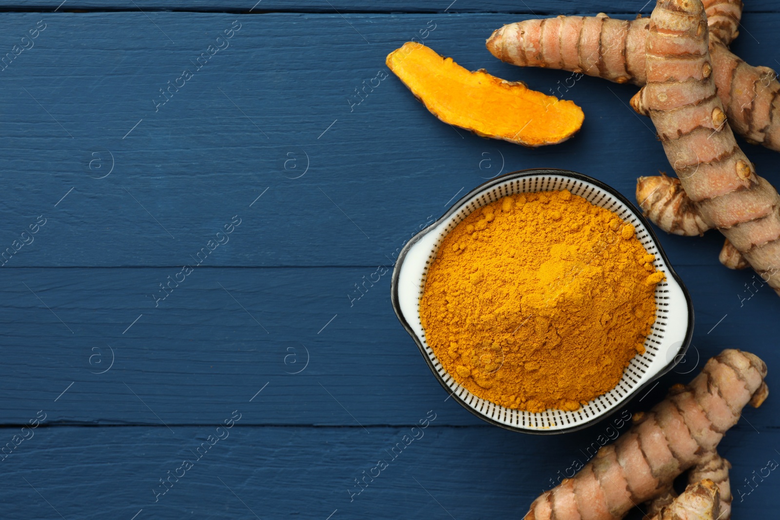 Photo of Bowl with turmeric powder and raw roots on blue wooden table, flat lay. Space for text