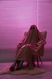 Glamorous ghost. Woman in sheet and high heel shoes on armchair in pink light